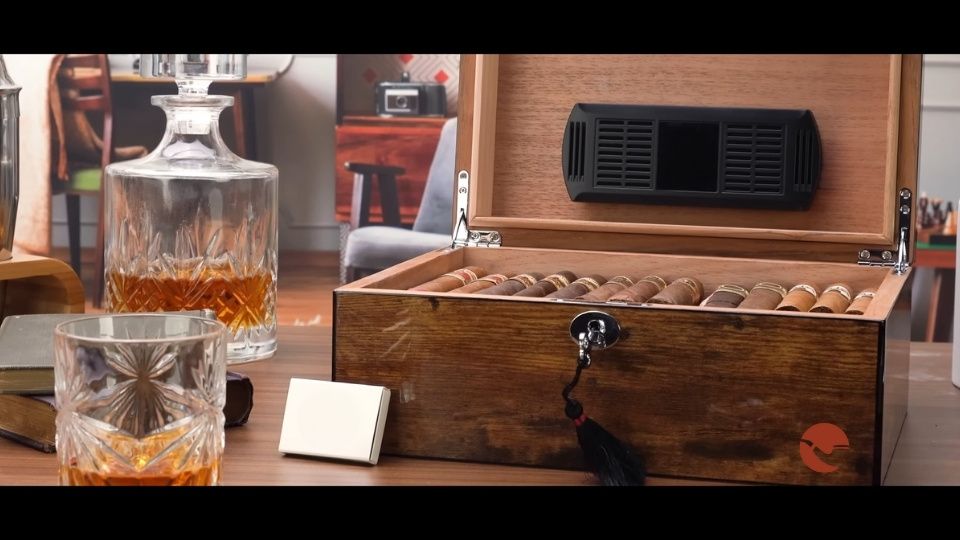 Open wooden humidor filled with cigars, placed beside whiskey glasses and a decanter on a wooden table.